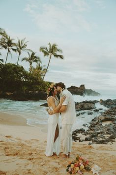 a man and woman standing on top of a sandy beach next to the ocean with palm trees