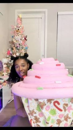 a woman holding up a giant pink cupcake in front of a christmas tree with decorations on it