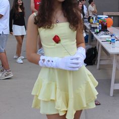 a woman in a yellow dress and white gloves holding a red rose while standing next to a table full of people