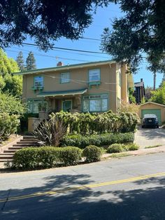 a yellow house with green trim and bushes on the front yard, next to a tree lined street