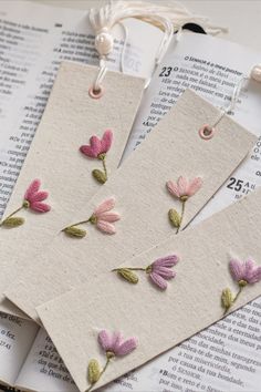 two bookmarks with flowers on them sitting on top of an open book next to thread