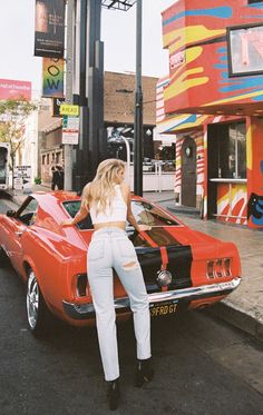 a woman leaning on the hood of a red car in front of a colorful building