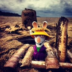 a stuffed animal wearing a yellow hat and purple dress sitting in the sand near logs