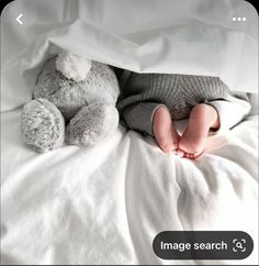 a baby laying under a blanket next to a teddy bear on top of a bed