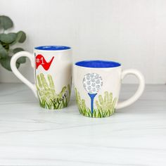 two coffee mugs sitting on top of a white counter next to a potted plant