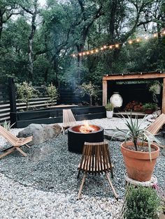 a fire pit surrounded by wooden chairs and lights in a backyard area with rocks, gravel, and trees