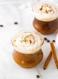 two mugs filled with hot chocolate and topped with whipped cream, cinnamon sticks, and powdered sugar