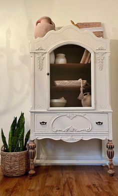 a white china cabinet sitting on top of a wooden floor next to a potted plant