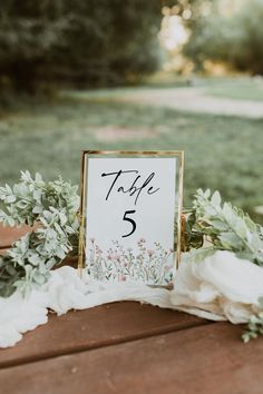 a table number sign sitting on top of a wooden table next to flowers and greenery