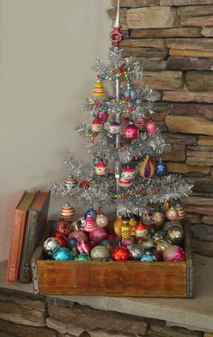 a small christmas tree with ornaments in a wooden crate next to a stone fireplace mantel