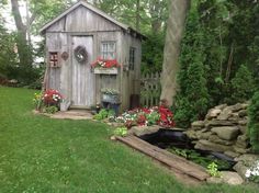 a garden shed is surrounded by flowers and rocks in the yard, with a pond