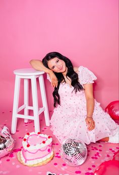 a woman sitting in front of a cake on top of a pink table with confetti