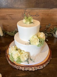 a three tiered cake with white flowers and greenery sits on a wooden platter