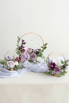 purple flowers and greenery are arranged on a white table cloth with gold hoop holders