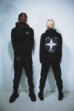 two people standing next to each other in front of a white wall wearing black clothing