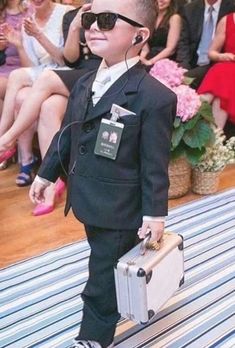 a young boy wearing sunglasses and carrying a suit case while standing on a rug with people in the background