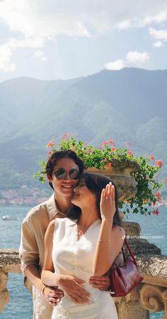 a man and woman standing next to each other in front of water with mountains behind them