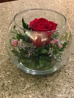 a red rose in a glass bowl filled with greenery and candles on a counter