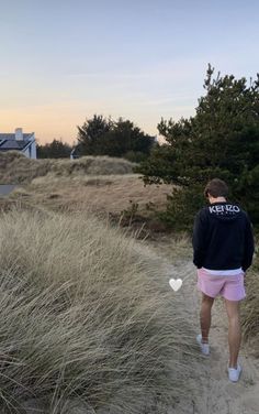 a man walking down a dirt path next to tall grass and trees at the beach