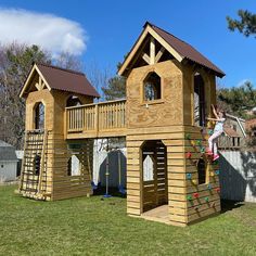 a child climbing on the top of a wooden play structure in a yard with grass and trees