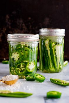 two jars filled with pickles and seasoning next to green peppers on a table