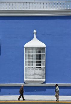 two people walking past a blue building with a white birdcage on it's side