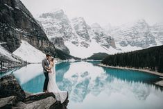 a bride and groom standing on the edge of a cliff overlooking a lake with snow covered mountains in the background