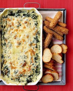 a casserole dish with spinach and cheese next to bread sticks on a red table