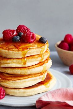 a stack of pancakes topped with berries and syrup on a white plate next to a bowl of raspberries