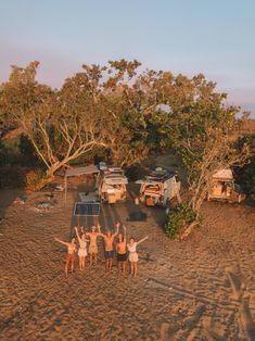 four people standing on the beach with their arms in the air and two campers behind them