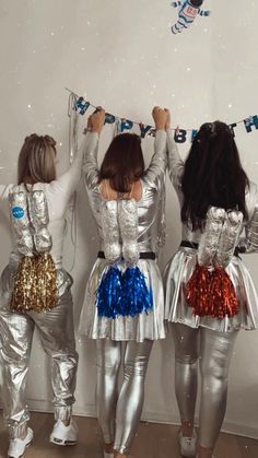 three girls dressed in silver and blue are holding their hands up above their heads as they stand against a white wall