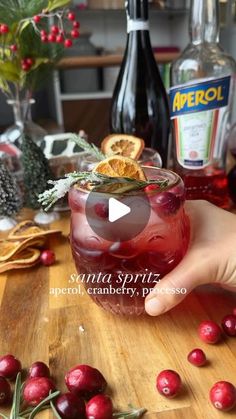 a person is holding a glass filled with cranberry shrub and oranges on the table