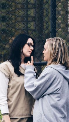 two women standing next to each other in front of a fence and looking at each other