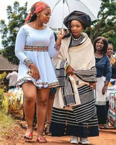 two women standing next to each other with an umbrella in their hands and people behind them