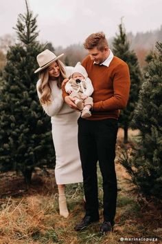 a man and woman holding a baby in their arms while standing next to christmas trees