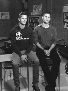 two young men sitting next to each other in front of desks and bookshelves