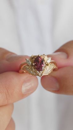 a person holding a gold ring with a red stone in it's center and leaves around the band