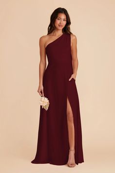 a woman in a burgundy one shoulder gown with thigh high slits, holding a white clutch