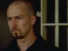 a man with a bald head and blue eyes stares into the distance while standing in front of a jail cell