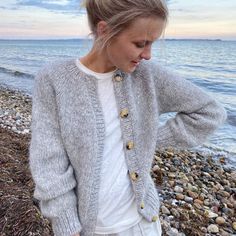 a young woman standing on top of a rocky beach next to the ocean wearing a gray cardigan