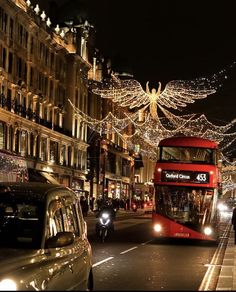 a red double decker bus driving down a street next to tall buildings covered in christmas lights