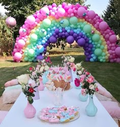 a table topped with lots of balloons and flowers next to a rainbow shaped cake on top of a white table