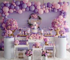 a table topped with lots of pink and purple flowers next to a wall covered in balloons