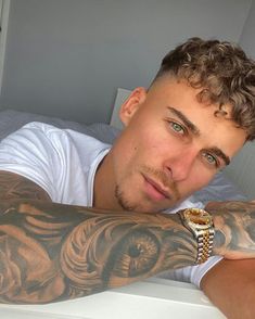 a man with tattoos and a watch on his arm is laying in bed looking at the camera