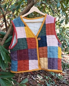 a multicolored vest hanging on a tree branch with leaves around it and another patchwork item in the background