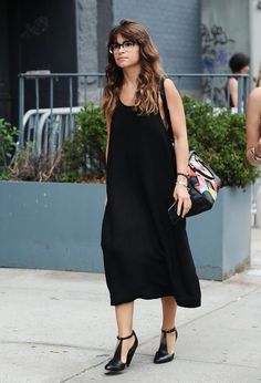 a woman in black dress walking down the street with her hand on her hip bag