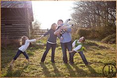 three adults and two children are playing with a dog in the grass near a barn