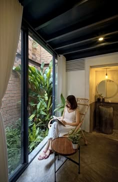 a woman sitting on a chair reading a book in front of a large window with plants outside