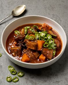 a white bowl filled with meat and vegetables on top of a gray table next to green peppers