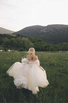 a woman in a white dress is walking through the grass with mountains in the background